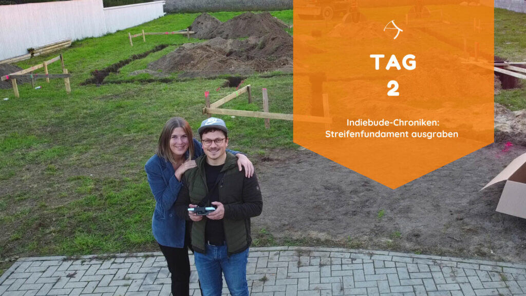 Am Tag 2 wird der Graben für das Streifenfundament ausgehoben. Sarah und Thomas stehen vor dem vorbereiteten Grundstück. Im Hintergrund sind Radbagger, Aushub der Fundamentegrube. Dreicksböcke und Snurgerüst.